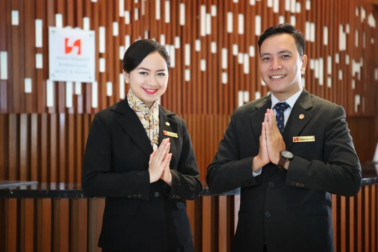 Two hotel staff members in professional uniforms greet guests with a traditional gesture, reflecting Indonesia's hospitality industry.