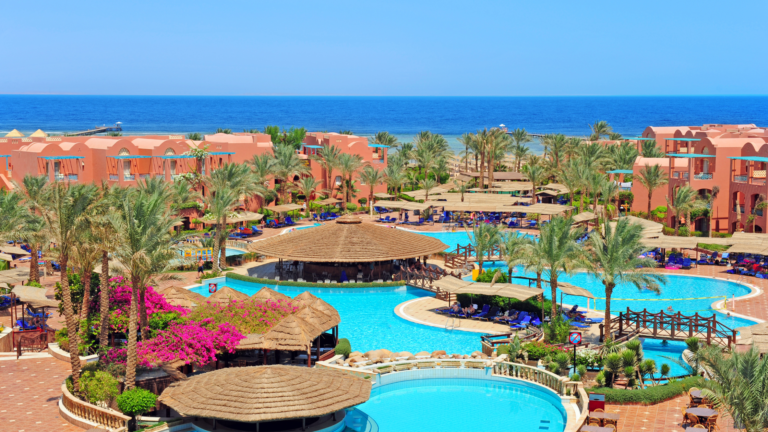 A luxurious seaside resort with multiple swimming pools, palm trees, and terracotta-colored buildings overlooking the ocean. The resort features thatched-roof cabanas, lounge chairs, and scenic landscaping, highlighting the potential for investment in resorts as a lucrative opportunity in the hospitality sector.