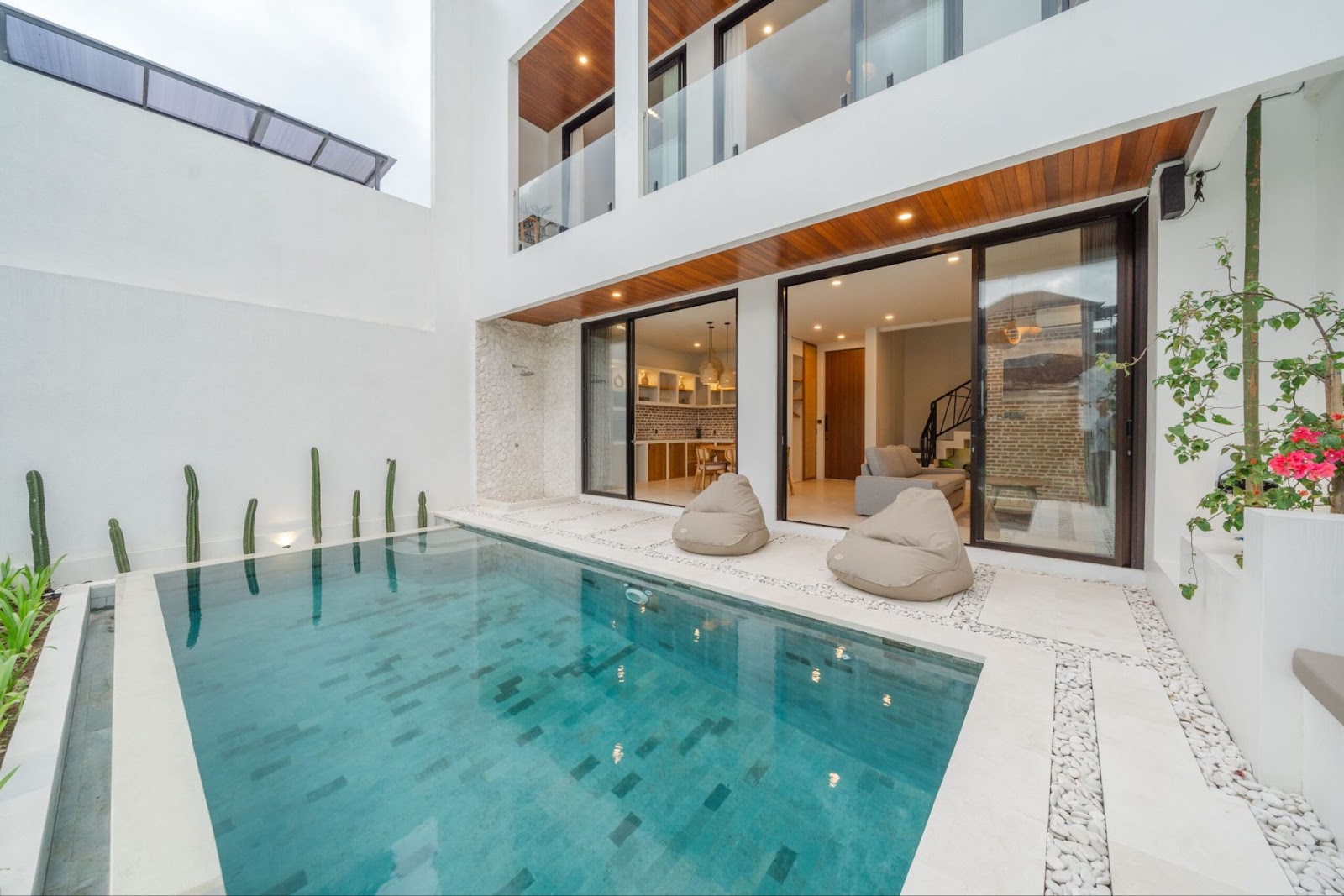 The contemporary exterior of a modern tropical Canggu Bali villa, showcasing a clean-lined swimming pool with beanbag chairs and large glass doors leading to the interior.