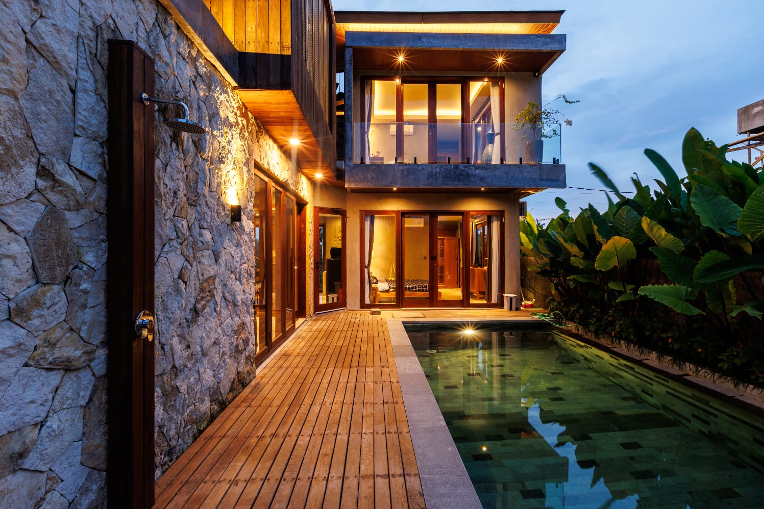 An elegant Villa, showcasing a natural stone wall with an outdoor shower, a wooden deck, and a swimming pool, all illuminated at dusk.