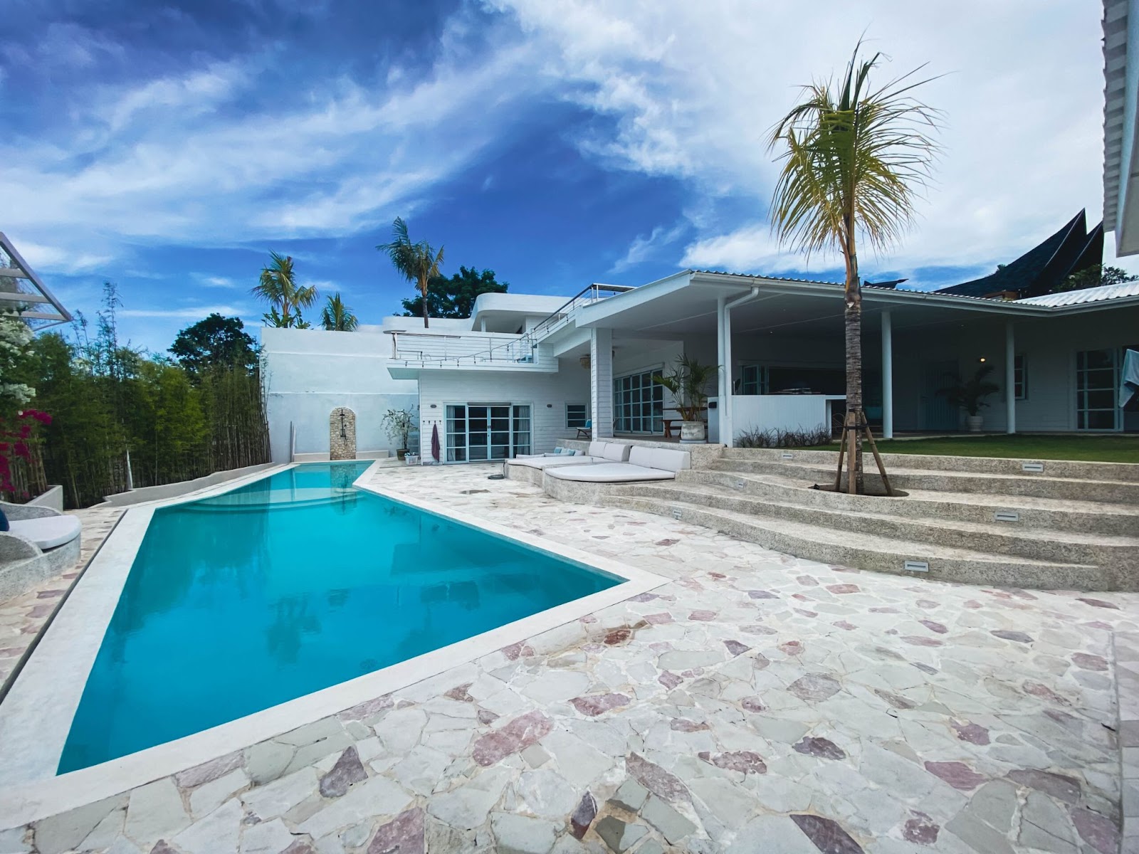 A beautiful 4-bedroom villa, with a large, bright blue swimming pool, expansive stone patio, and palm trees under a sunny sky.