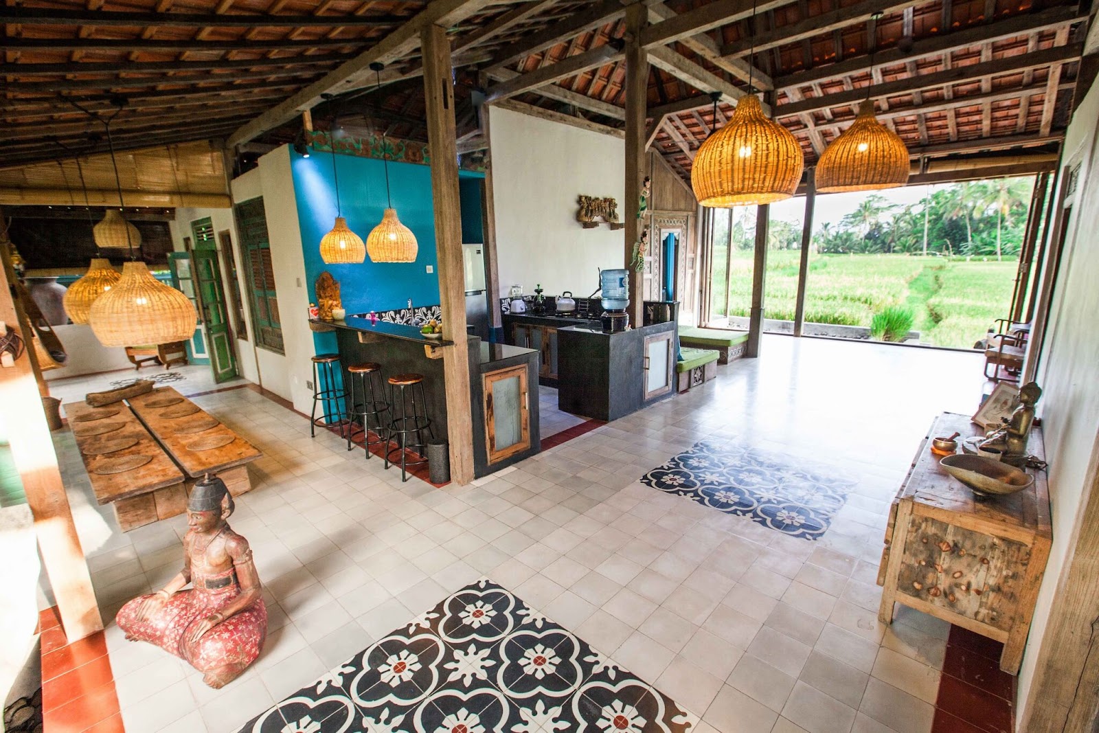 A spacious Joglo-style villa in Ubud with an open-plan living area, kitchen, and dining space, featuring traditional decor, woven pendant lights, and large windows offering panoramic views of lush green rice fields.