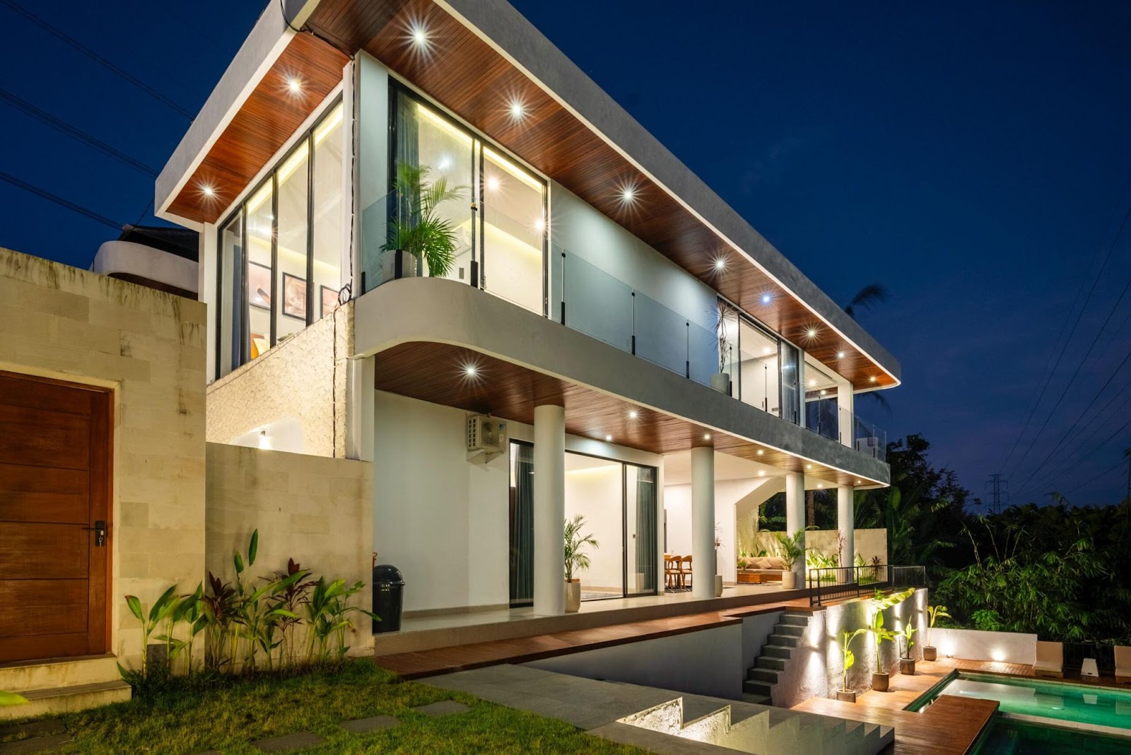 A modern, lit-up Villa, featuring large glass windows, a multi-level design, and steps leading down to a pool area.