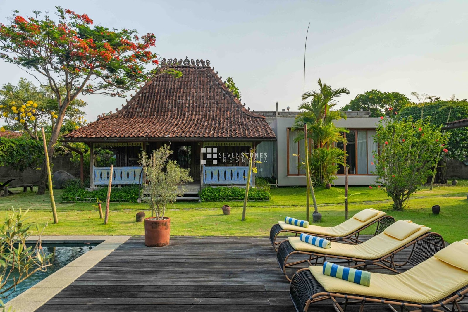 A bright Bali villa design features a traditional pavilion with a terracotta roof overlooking a wooden deck, lounge chairs, a pool, and a vibrant tropical garden under a clear sky.
