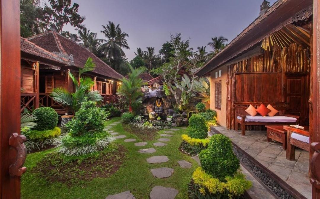 A tranquil Bali villa design showcases a stone pathway winding through lush gardens with traditional wooden architecture visible at dusk.