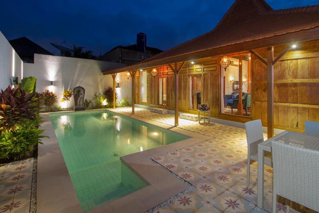 An illuminated Bali villa design at dusk displays a private pool bordered by ornate tiling and traditional wooden architecture with inviting interiors.