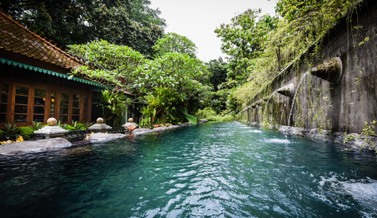 A lush, tropical swimming pool at an eco-friendly villa for sale in Bali, featuring a traditional building and a wall with water spouts surrounded by vibrant greenery.