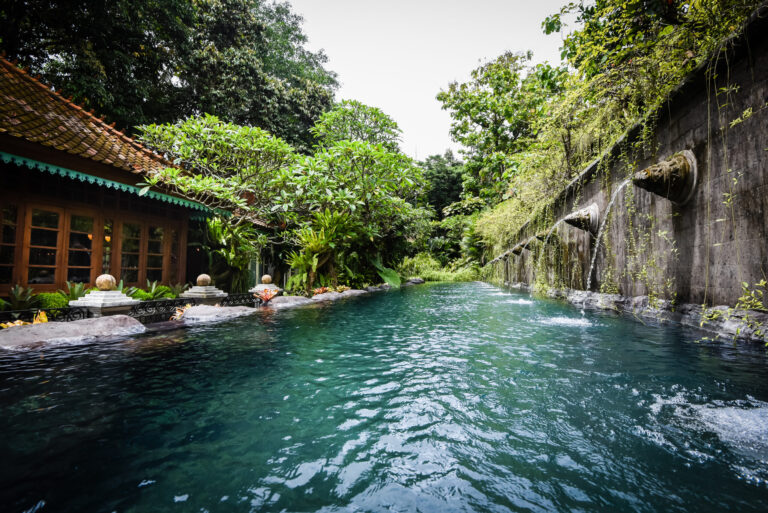 A lush, tropical swimming pool at an eco-friendly villa for sale in Bali, featuring a traditional building and a wall with water spouts surrounded by vibrant greenery.