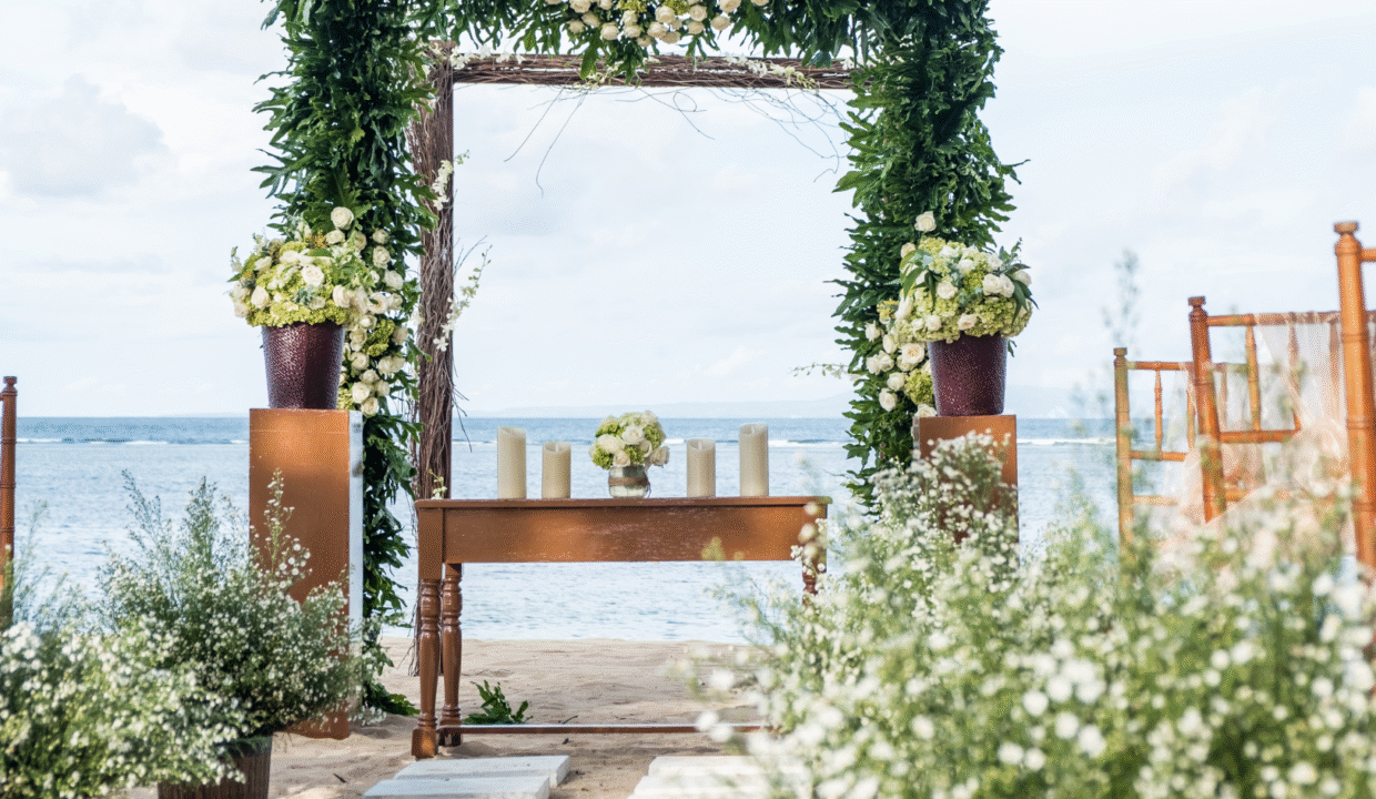 A beautifully decorated Bali wedding venue on the beach, featuring a floral arch, wooden table with candles, and ocean views in the background.
