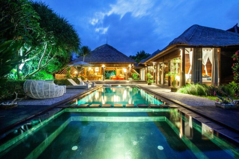 A serene Bali villa design features a central swimming pool reflecting the twilight sky and softly lit, open-air living spaces with thatched roofs, surrounded by lush tropical greenery.