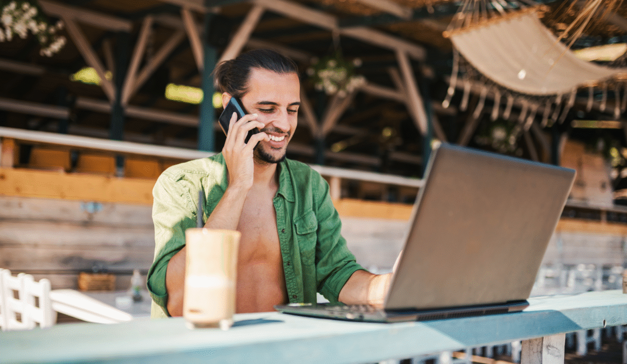A digital nomad working on a laptop at a beachside café while talking on the phone, representing Bali villas for digital nomads and the remote-work lifestyle.