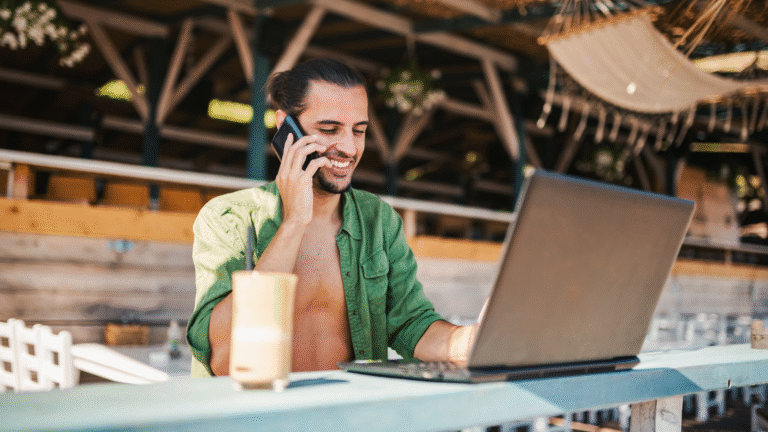 A digital nomad working on a laptop at a beachside café while talking on the phone, representing Bali villas for digital nomads and the remote-work lifestyle.