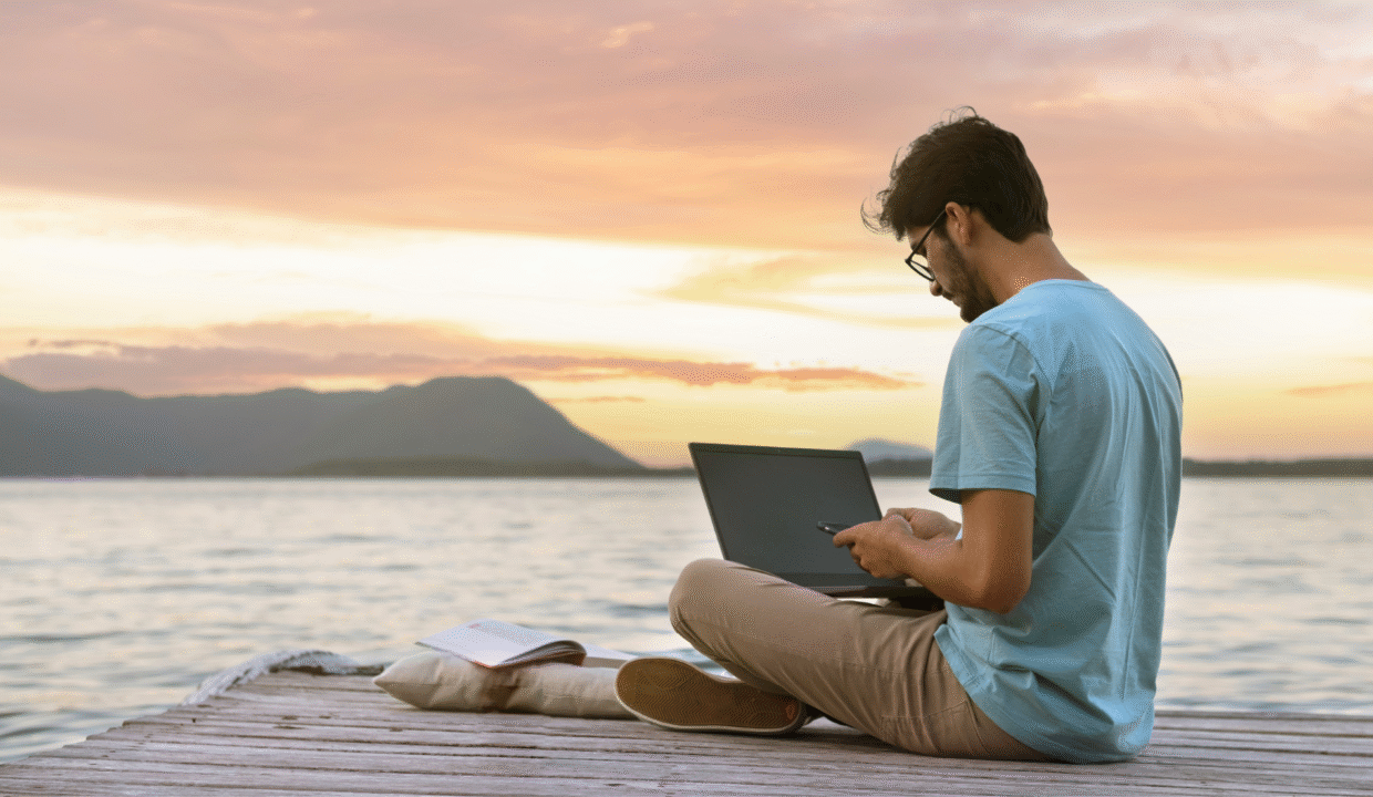 A man working remotely in Indonesia on his laptop outside with ocean and mountain views.