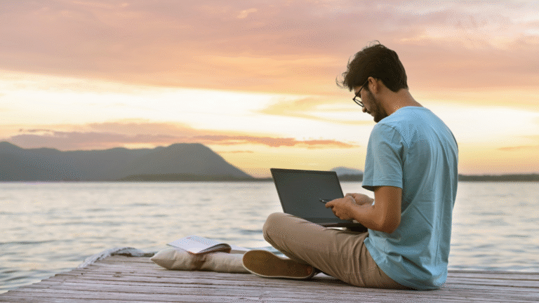 A man working remotely in Indonesia on his laptop outside with ocean and mountain views.